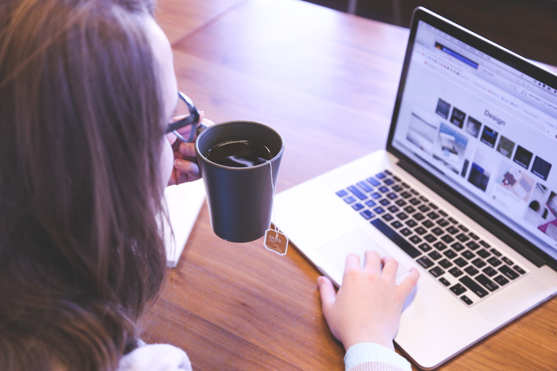 A woman sitting in front of an open laptop, holding a cup of tea in her left hand and scrolling on the mousepad with her right hand.