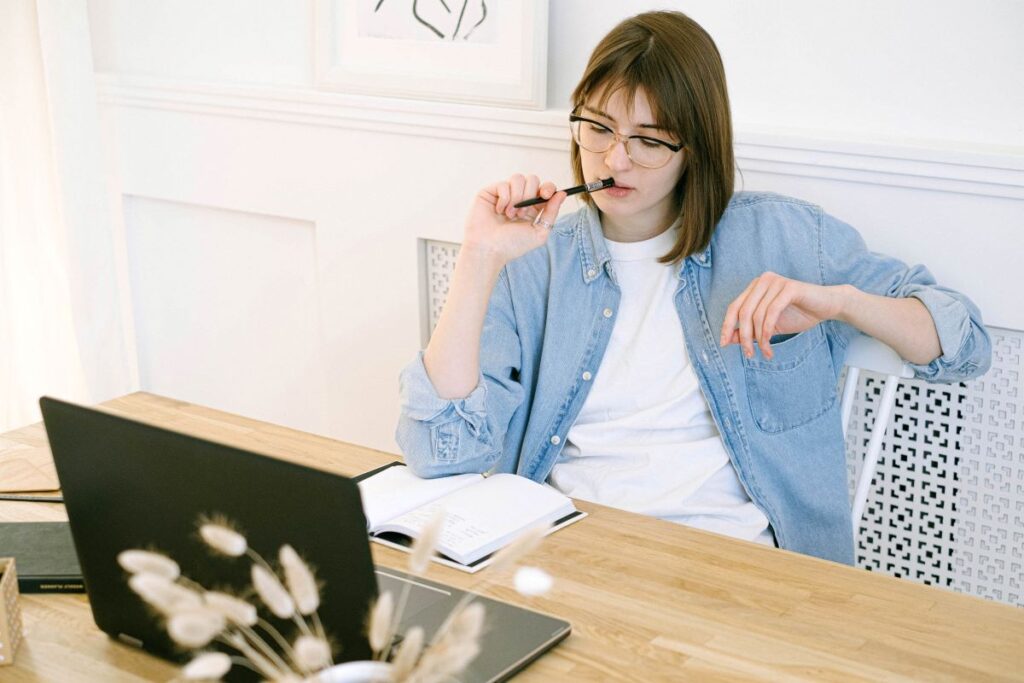 Woman sitting at her desk holding a pen against her mouth while looking thoughtfully at her laptop.