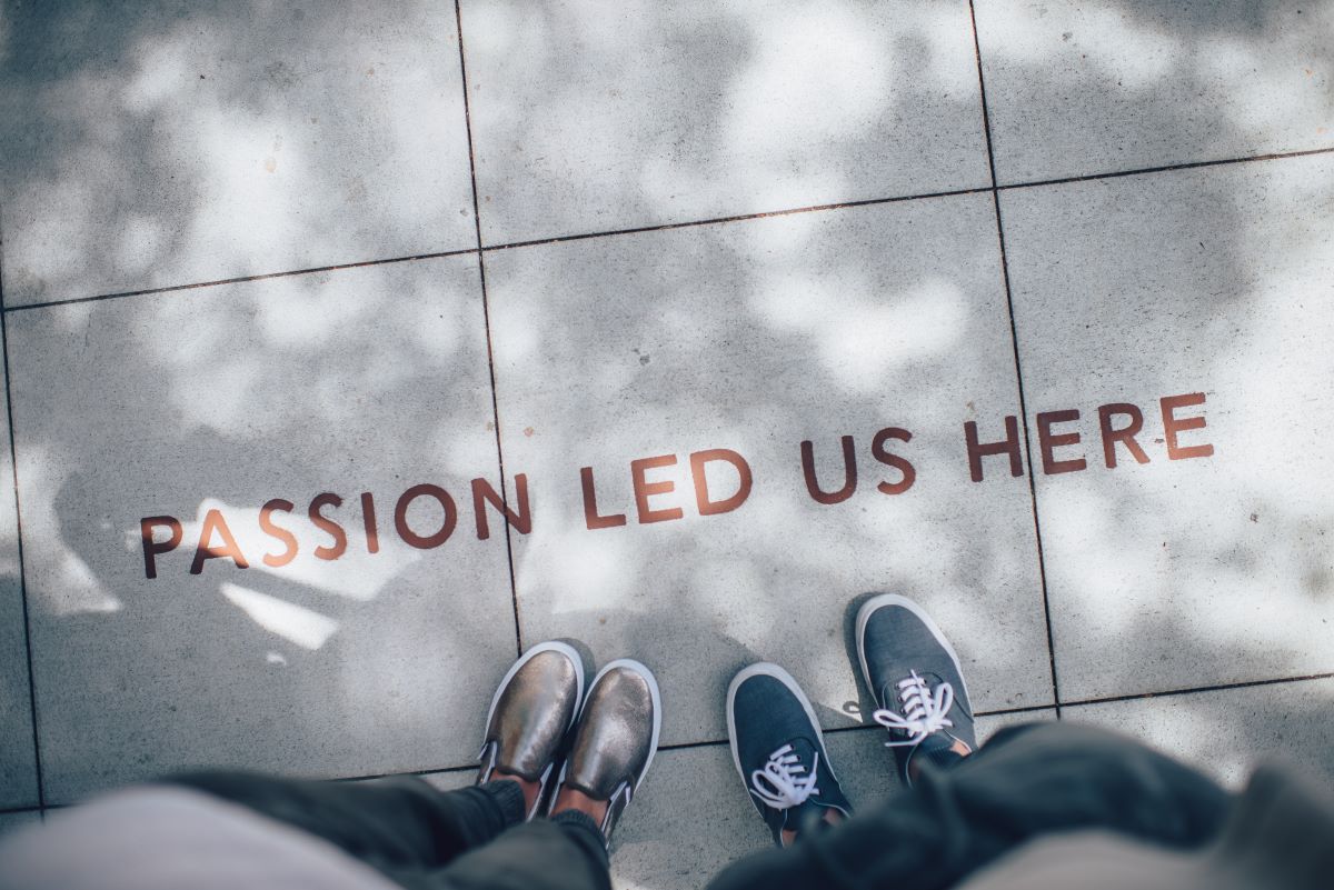 A view from above showing two people's feet standing on a pavement in front of the words 'Passion led us here' written in red capital letters on the pavement.