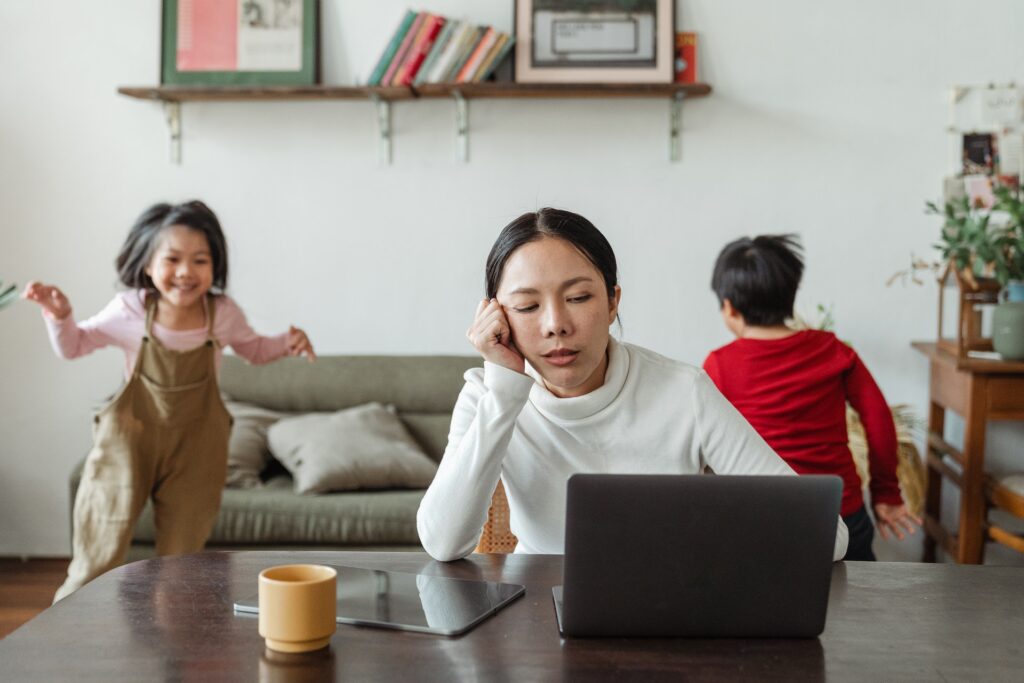 A mum sitting in front of her laptop in the lounge, with her head on her hand, looking exhausted, with her two young kids running around and playing behind her
