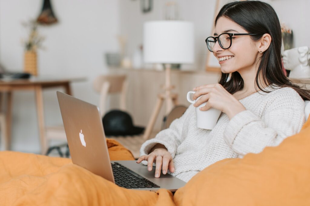 A woman, grinning and holding a mug of tea, is sitting on a sofa with an open laptop in front of her