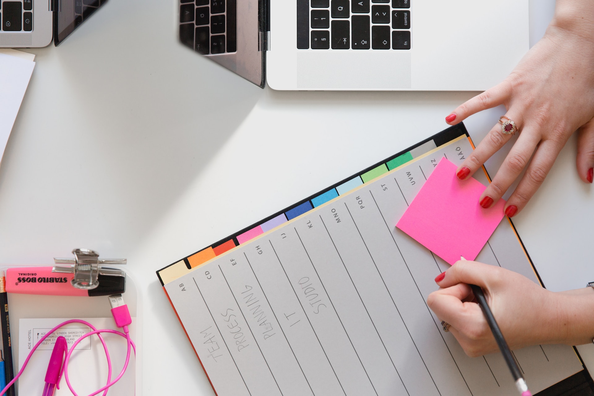 An open file of colourful dividers on a white desk - a woman's right hand is holding a pink Post-It note on top of the dividers and she is about to start writing on it using the pencil that's in her other hand