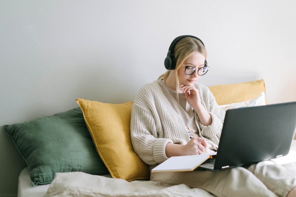 A woman sitting propped against large cushions, wearing headphones and writing in a notebook that's open on top of an open laptop that's on her lap