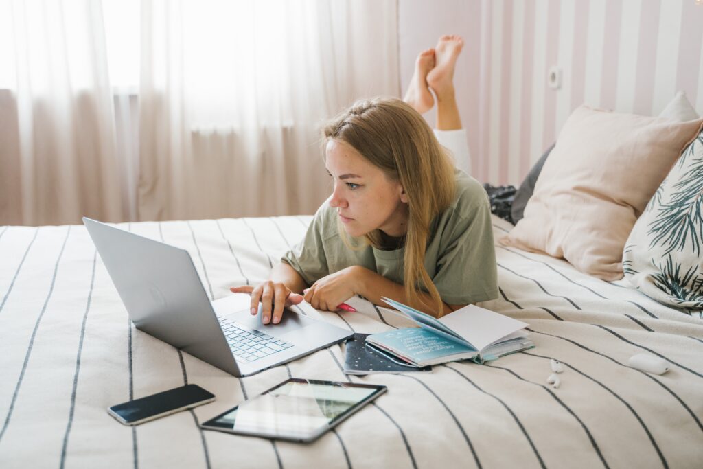 A woman lying on a bed with her feet up in the air behind her, typing on an open laptop, with a tablet, mobile phone and books next to her on the bed