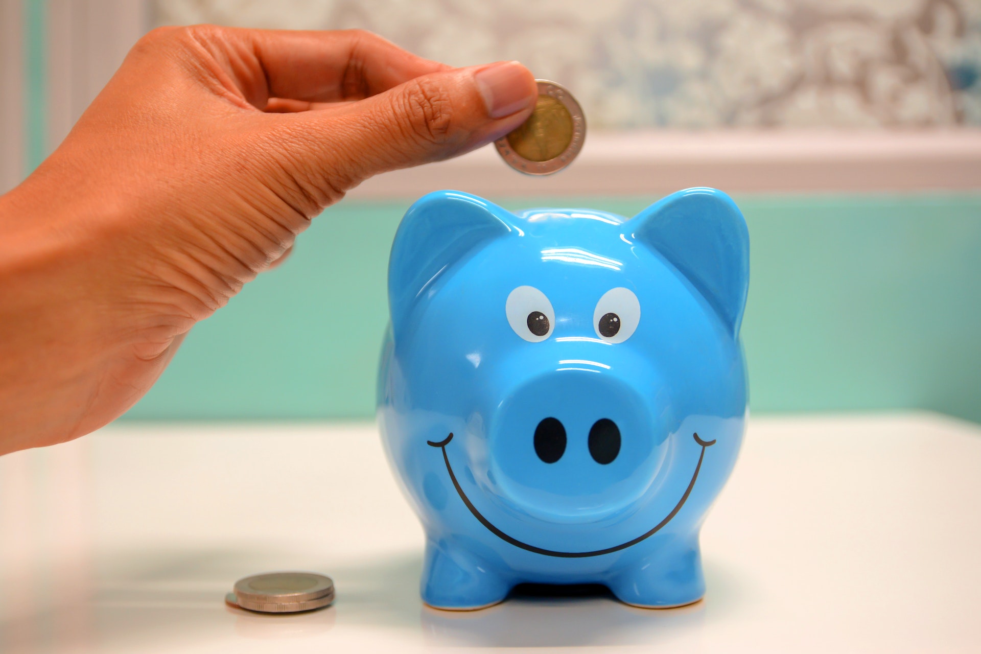 A smiling blue piggy bank with two coins next to it and a woman's hand dropping a coin into the top of it