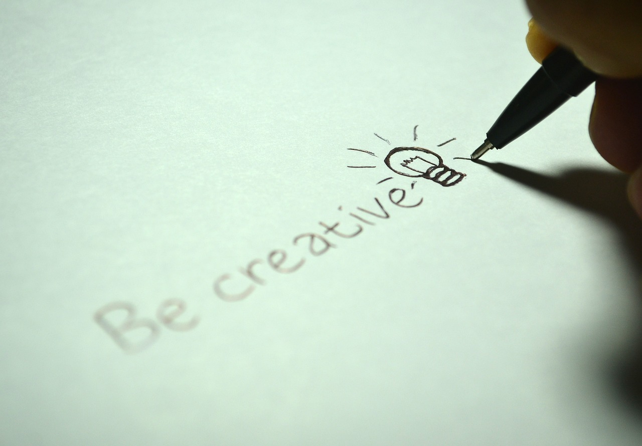 A hand holding a black biro, writing the words 'Be creative' and a picture of a light bulb
