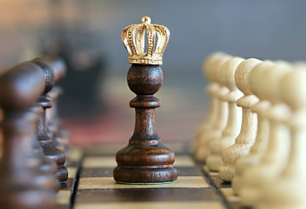 A mahogany pawn wearing a gold crown standing on a chess board with a row of white pawns to the right of it and a row of mahogany pawns to the left of it