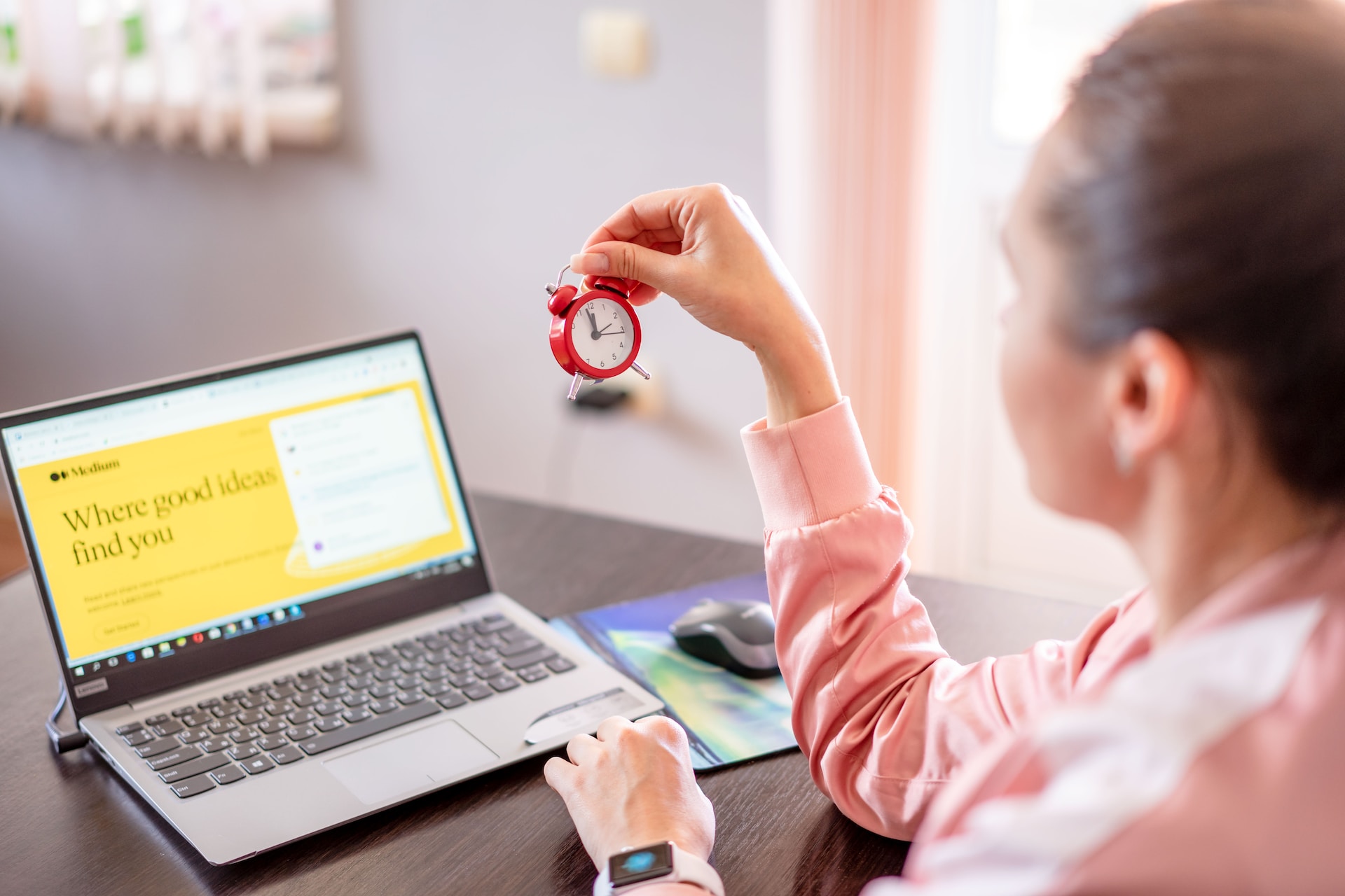 A woman sitting at a desk with her laptop open in front of her, holding a mini alarm clock in her right hand