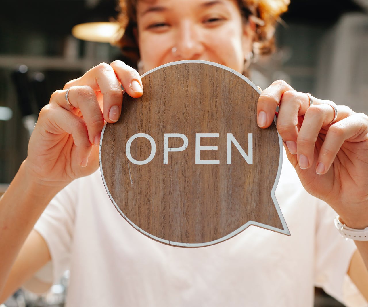 A woman holding a wooden speech bubble with the word 'OPEN' written on it in white ink.