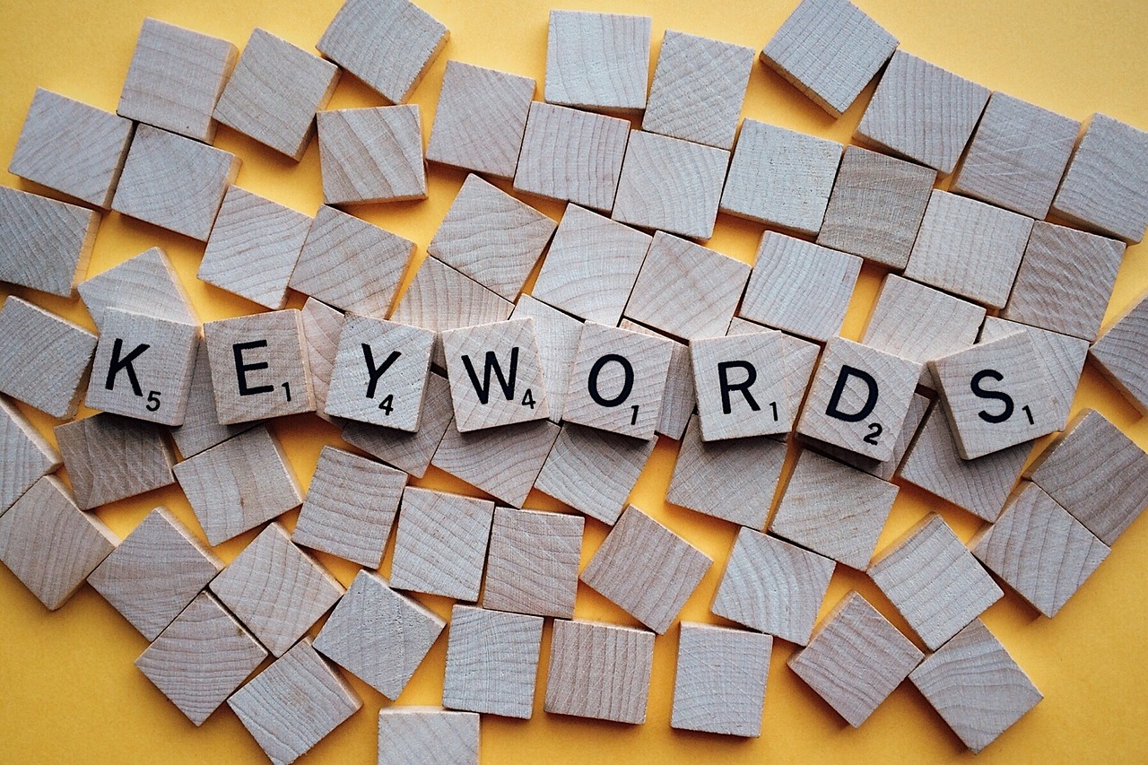 The word 'KEYWORDS' spelled out on wooden Scrabble tiles that are on top of a pile of blank tiles placed on a yellow surface.