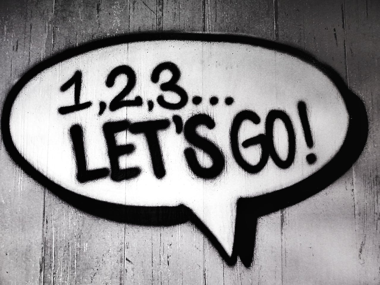 A black and white image of a graffiti speech bubble on wooden planks with '1,2,3...LET'S GO!' written inside it.