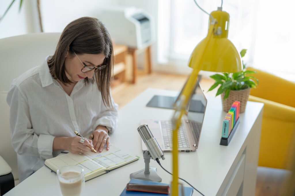 A woman is sitting at a desk with an open laptop, a microphone and a desk lamp in front of her. She is writing in a diary.