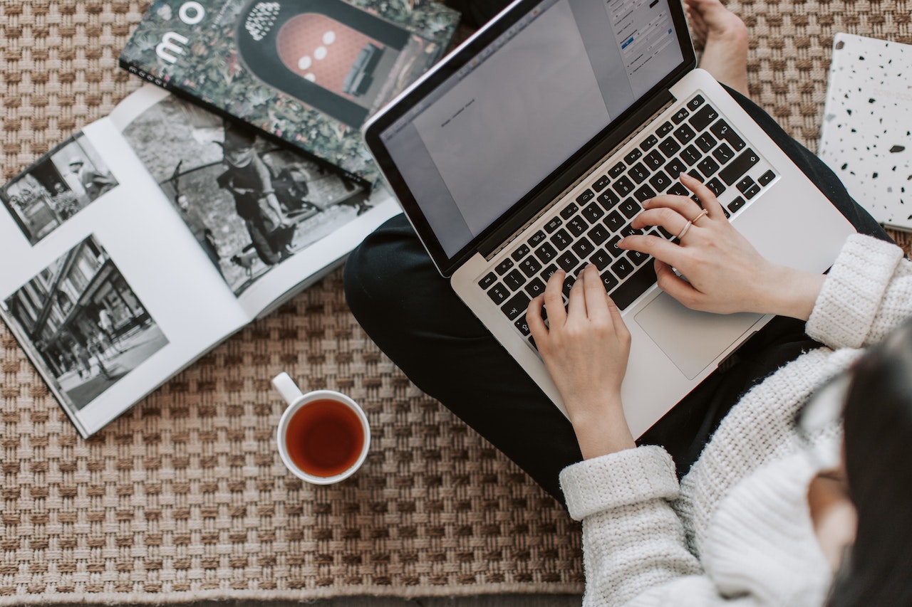 An aerial view of a woman sitting on a rug and typing on a laptop, with a cup of tea and some books next to her.