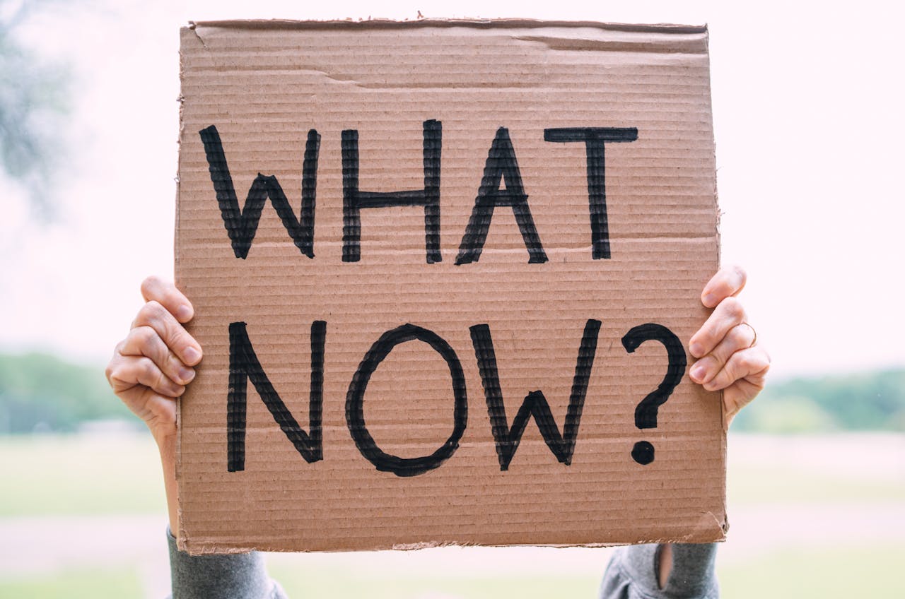 A cardboard sign being held up by a woman above her head with the words 'WHAT NOW? written in thick black ink.