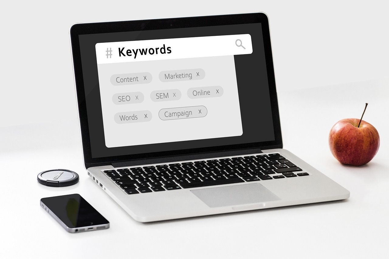 An open laptop with the word 'Keywords' displayed at the top and various keywords relating to SEO underneath it. To the right of the laptop is a red apple and to the left of it is a mobile phone.