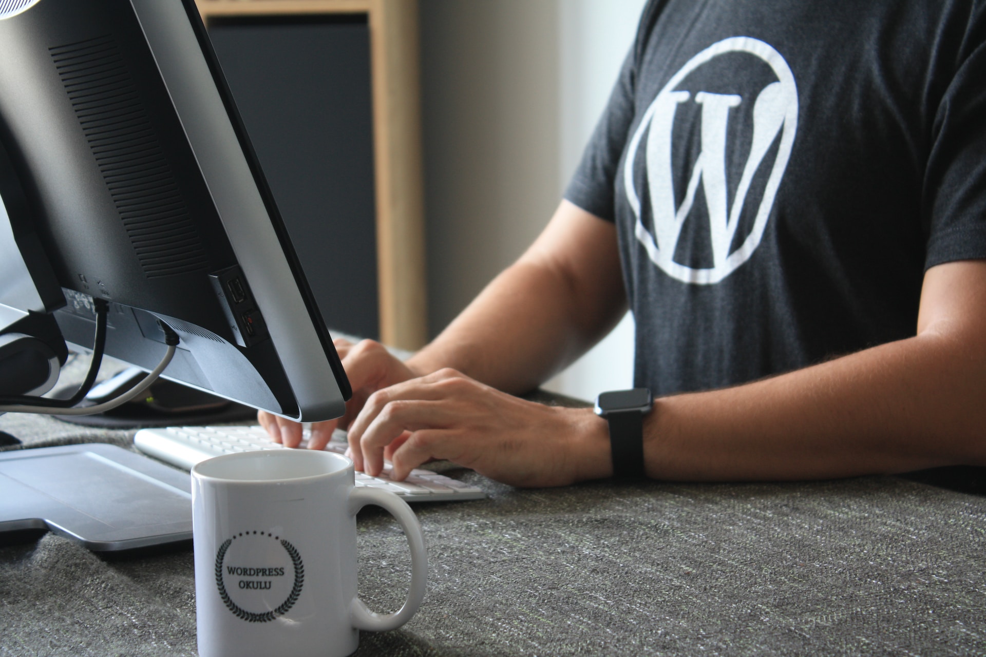 A cut-off shot of a person wearing a T-shirt with the WordPress logo on it, sitting in front of a computer and typing on the keyboard