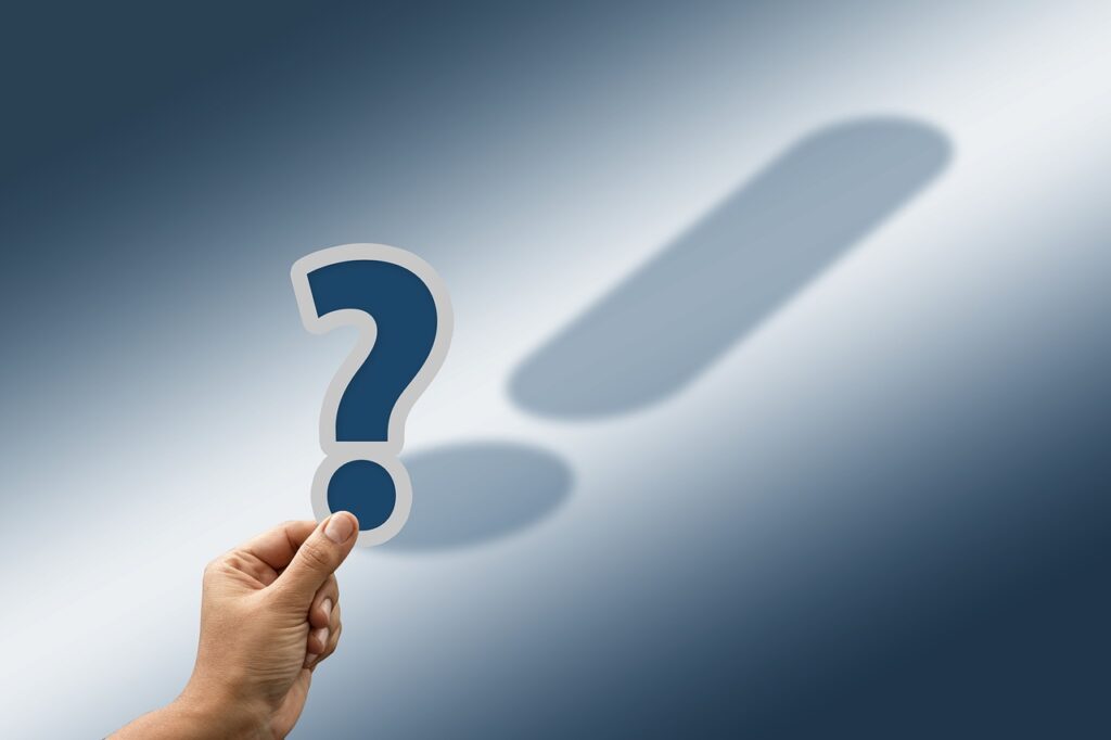 A hand is holding up a cut-out blue question mark with a white border. The shadow of the question mark is actually an exclamation mark.