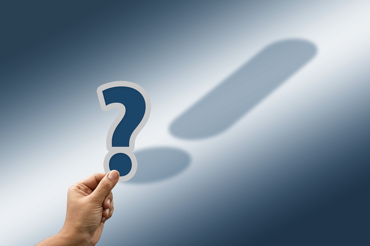 A hand is holding up a cut-out blue question mark with a white border. The shadow of the question mark is actually an exclamation mark.