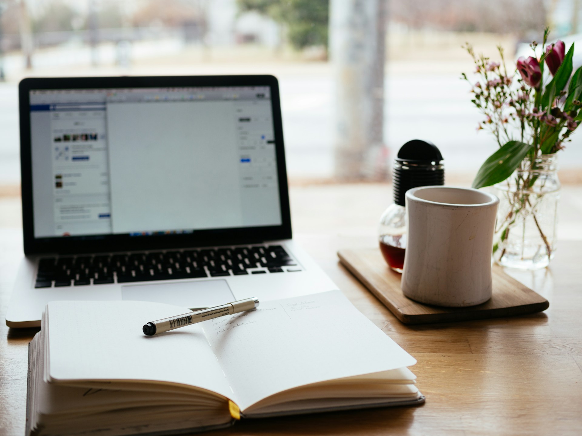 An open notebook with a pen resting across the pages, in front of an open laptop, with a mug and a vase of flowers next to them.