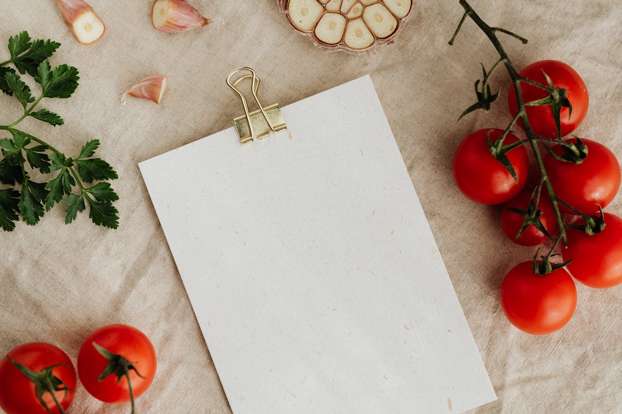 A blank piece of parchment paper with a gold bulldog clip at the top, on a natural-coloured cloth with vine cherry tomatoes, garlic cloves and a sprig of parsley leaves placed around the paper.