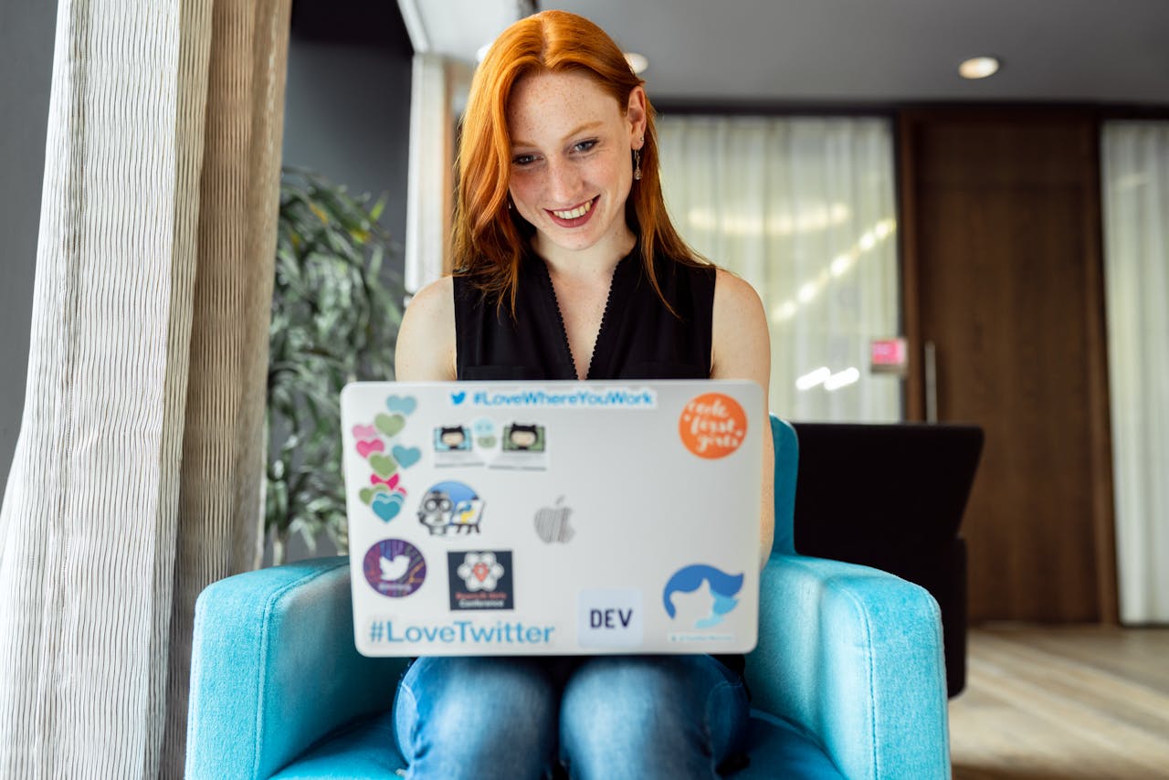 A red-haired woman is sitting in a blue armchair, grinning while she's looking at her laptop, which is on her lap and has colourful stickers on its cover.